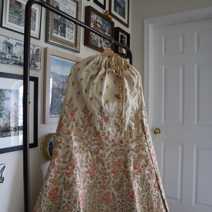 May include: A cream-colored skirt with gold and pink embroidery. The skirt has a drawstring waist and is hanging on a black clothing rack.