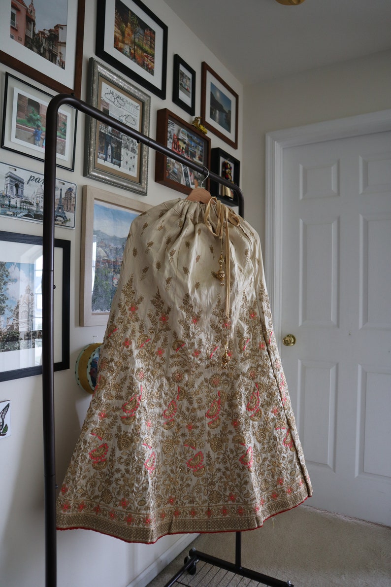 May include: A cream-colored skirt with gold and pink embroidery. The skirt has a drawstring waist and is hanging on a black clothing rack.