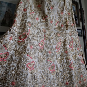 May include: A white skirt with gold and pink embroidery. The skirt is made of a soft, flowing fabric and has a delicate floral pattern.