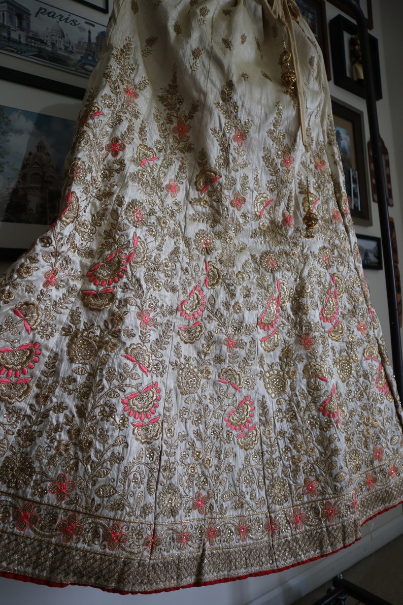 May include: A white skirt with gold and pink embroidery. The skirt is made of a soft, flowing fabric and has a delicate floral pattern.
