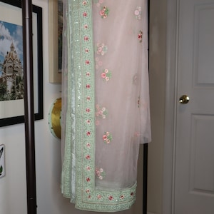 May include: A light pink sheer scarf with a green embroidered border and floral designs. The scarf is hanging on a wooden hanger.