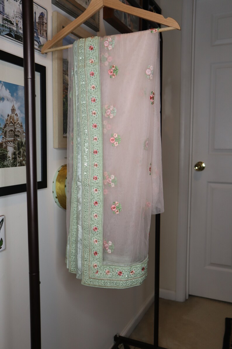 May include: A light pink sheer scarf with a green embroidered border and floral designs. The scarf is hanging on a wooden hanger.