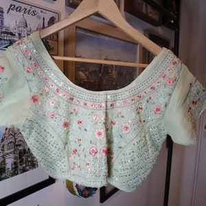 May include: A light green embroidered blouse with pink floral details and sequins. The blouse has a scoop neckline and short sleeves.