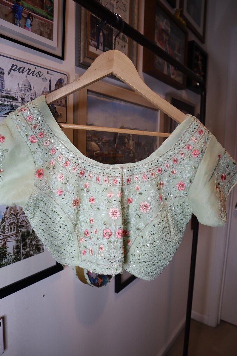 May include: A light green embroidered blouse with pink floral details and sequins. The blouse has a scoop neckline and short sleeves.