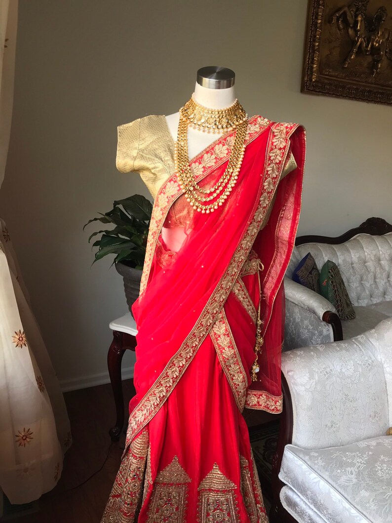 Op de afbeelding: Een rode en gouden Indiase bruidsoutfit met ingewikkelde borduursels en versieringen. De outfit bestaat uit een lange rok, een doorschijnende dupatta en een bijpassend blouse. De outfit is versierd met gouden sieraden, waaronder een ketting, oorbellen en armbanden.