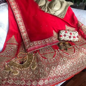 Op de afbeelding: Een rode en gouden geborduurde bruidslehenga met een bijpassend gouden blouse en een kleine clutch. De lehenga heeft ingewikkelde bloem- en geometrische patronen en is versierd met pailletten en kralen.