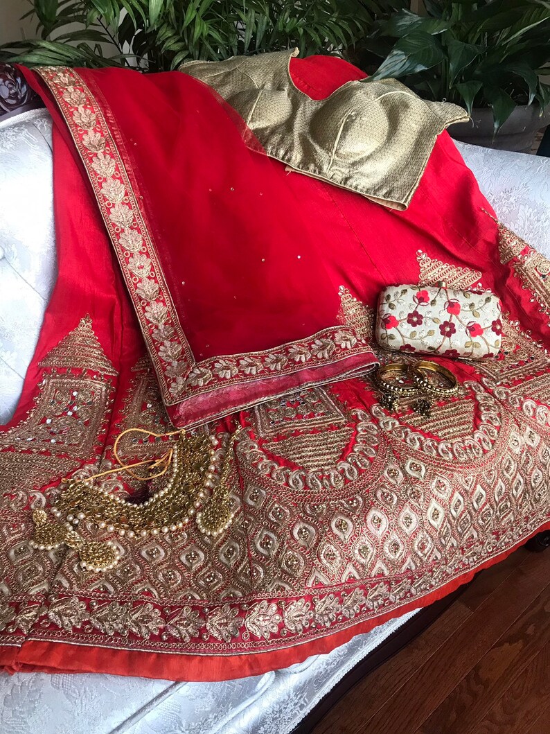 Op de afbeelding: Een rode en gouden geborduurde bruidslehenga met een bijpassend gouden blouse en een kleine clutch. De lehenga heeft ingewikkelde bloem- en geometrische patronen en is versierd met pailletten en kralen.