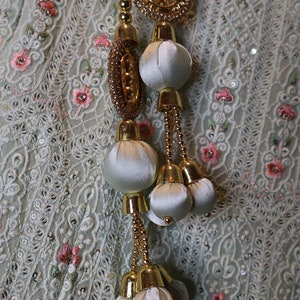 May include: A white fabric with gold and white embellishments. The embellishments are in the shape of round balls and circles. The fabric has a delicate pattern.