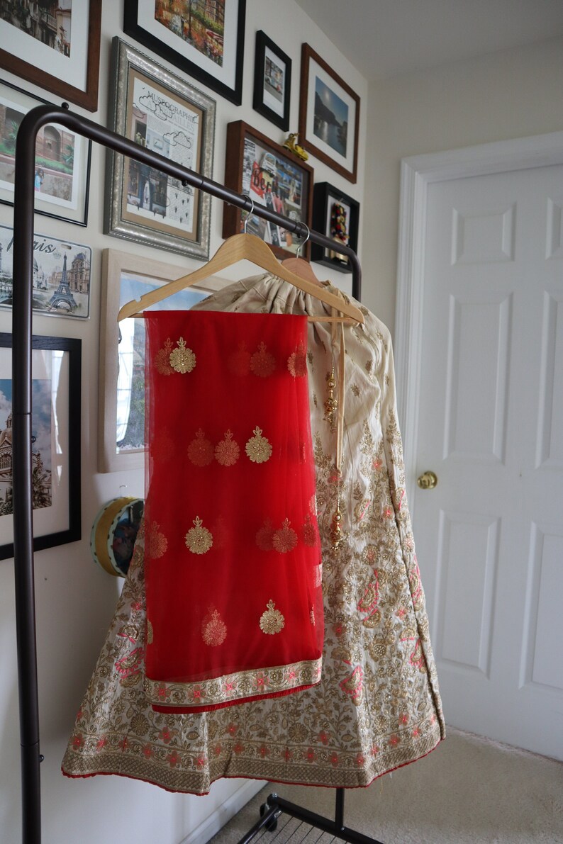 May include: A white and gold embroidered lehenga with a red sheer dupatta. The dupatta has gold embroidery and is draped over the lehenga.