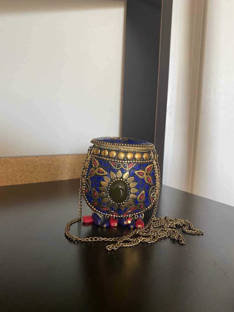 Puede incluir: Una peque&ntilde;a caja redonda de metal azul y dorado con una cadena y un borla. La caja est&aacute; decorada con intrincados patrones florales y una piedra preciosa verde.