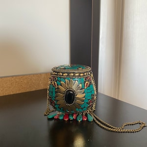 Puede incluir: Un peque&ntilde;o bolso de mano redondo de color turquesa y dorado con una piedra negra en el centro. El bolso tiene una correa de cadena y borlas rojas y turquesas.
