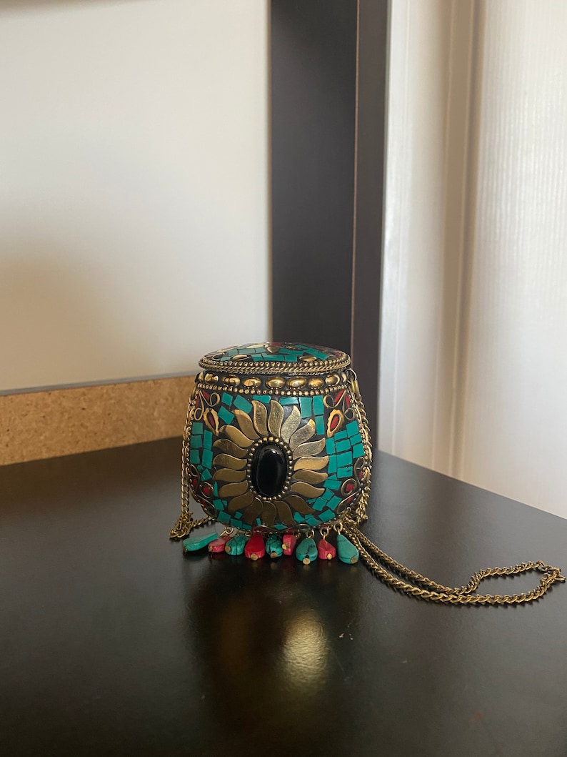 Puede incluir: Un peque&ntilde;o bolso de mano redondo de color turquesa y dorado con una piedra negra en el centro. El bolso tiene una correa de cadena y borlas rojas y turquesas.