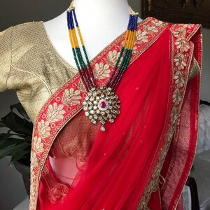 Op de afbeelding: Een rode en gouden sari met ingewikkelde borduursels en een veelkleurige kralenketting met een grote hanger.