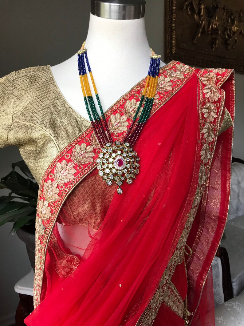 Op de afbeelding: Een rode en gouden sari met ingewikkelde borduursels en een veelkleurige kralenketting met een grote hanger.