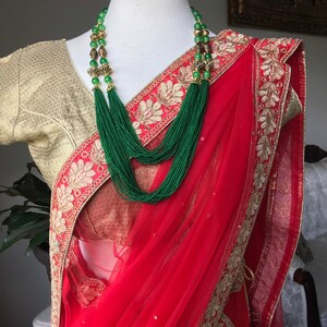 Op de afbeelding: Een rode en gouden sari met een groene kralenketting. De sari is gemaakt van een transparante stof met een gouden geborduurde rand. De ketting is gemaakt van groene kralen en gouden accenten.