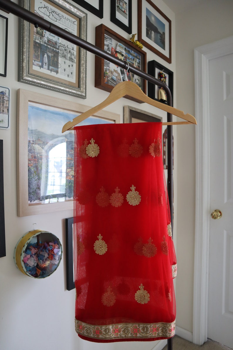 May include: A red sheer saree with gold embroidery and a gold and pink border. The saree is hanging on a wooden hanger.