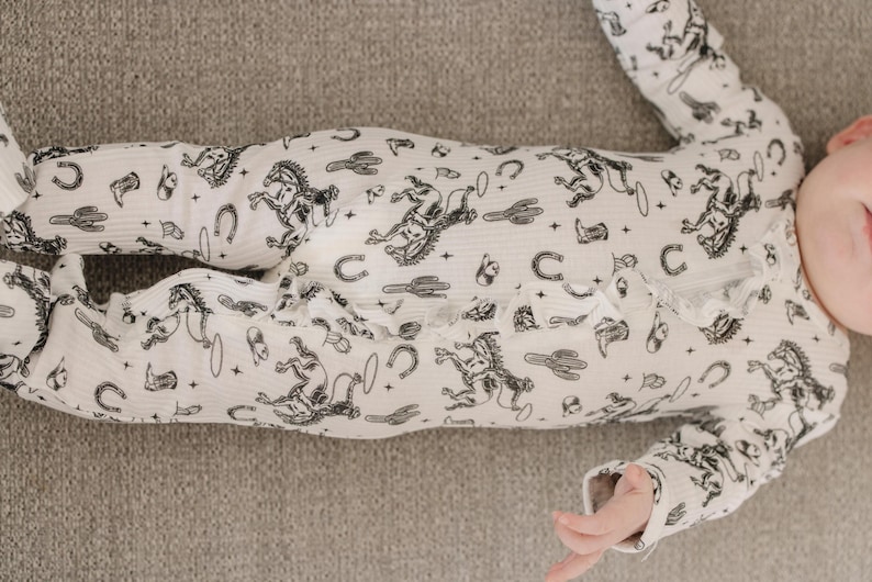 K&ouml;nnte beinhalten: Ein wei&szlig;er Baby-Strampler mit einem schwarz-wei&szlig;en Druck von Pferden, Kakteen und anderen Western-Motiven.