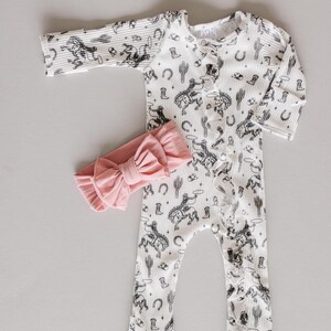 K&ouml;nnte beinhalten: Ein wei&szlig;er Baby-Strampler mit einem schwarz-wei&szlig;en Cowboy-Druck. Der Strampler hat einen Knopfverschluss und lange &Auml;rmel. Ein rosafarbenes Schleifen-Stirnband ist im Lieferumfang enthalten.