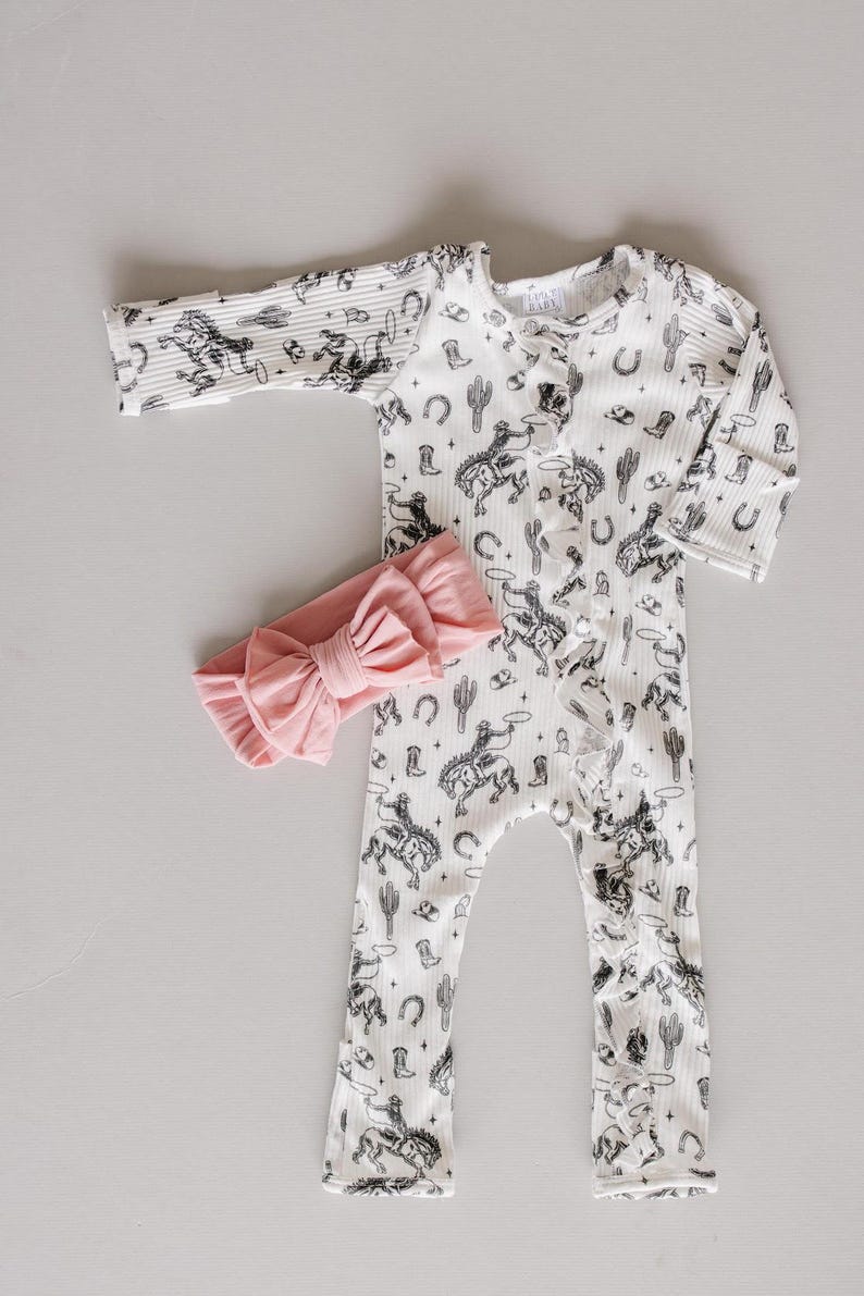 K&ouml;nnte beinhalten: Ein wei&szlig;er Baby-Strampler mit einem schwarz-wei&szlig;en Cowboy-Druck. Der Strampler hat einen Knopfverschluss und lange &Auml;rmel. Ein rosafarbenes Schleifen-Stirnband ist im Lieferumfang enthalten.