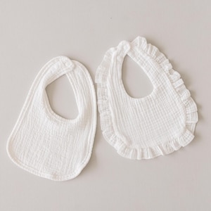 May include: Two white baby bibs, one with a ruffled edge and one without. Both bibs are made of a soft, textured fabric.
