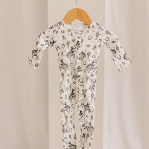 K&ouml;nnte beinhalten: Ein wei&szlig;er lang&auml;rmliger Baby-Strampler mit einem schwarz-wei&szlig;en Druck von Cowboys, Pferden, Kakteen und Sternen. Der Strampler hat einen Knopfverschluss an der Vorderseite.