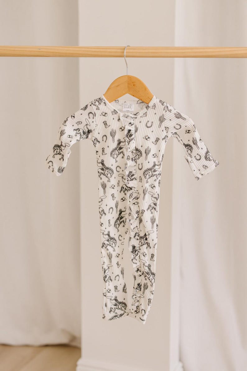 K&ouml;nnte beinhalten: Ein wei&szlig;er lang&auml;rmliger Baby-Strampler mit einem schwarz-wei&szlig;en Druck von Cowboys, Pferden, Kakteen und Sternen. Der Strampler hat einen Knopfverschluss an der Vorderseite.