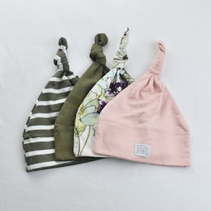May include: Four knotted baby hats in different patterns. One hat is striped in green and white, one has a floral pattern, one is solid green, and one is solid pink with a white label that says "Dulce Baby".