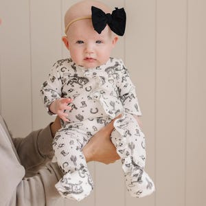 K&ouml;nnte beinhalten: Ein Baby tr&auml;gt einen wei&szlig;en und schwarzen lang&auml;rmeligen Strampler mit Western-Druck. Der Strampler hat R&uuml;schen an den &Auml;rmeln und Beinen. Das Baby tr&auml;gt ein schwarzes Schleifen-Haarband.