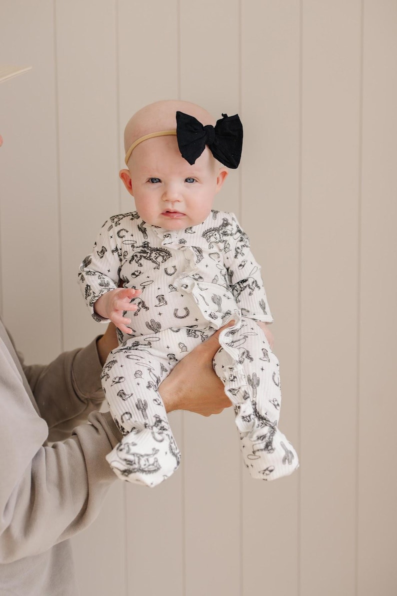 K&ouml;nnte beinhalten: Ein Baby tr&auml;gt einen wei&szlig;en und schwarzen lang&auml;rmeligen Strampler mit Western-Druck. Der Strampler hat R&uuml;schen an den &Auml;rmeln und Beinen. Das Baby tr&auml;gt ein schwarzes Schleifen-Haarband.
