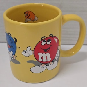 May include: A bright yellow ceramic mug featuring colorful M&M's character graphics on the exterior, including a red character and a blue character. The interior of the mug also features a small orange M&M's character printed near the rim.