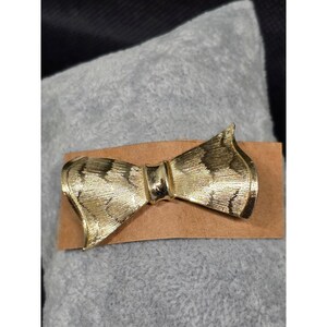 May include: A gold-toned metal brooch shaped like a bow. The surface of the bow features a textured, etched pattern with fine lines that create a subtle contrast against the polished, smooth center knot that connects the two flared sides.