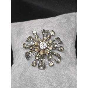 May include: A silver-toned metal brooch featuring a starburst design with multiple radiating ribbon-like loops. The center is set with a single clear faceted stone, while smaller clear stones are embedded at the tips of several outer loops.