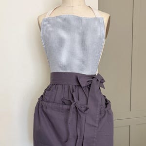 May include: An apron displayed on a mannequin. The top is blue and white striped, while the bottom is a solid dark purple. It features a wraparound design with a bow and pockets, and white straps.