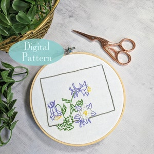 Puede incluir: Un bastidor de bordado blanco con un diseño floral en morado, amarillo y verde. El diseño es una representación estilizada de una flor dentro de un contorno gris de un estado. El texto "Digital Pattern" está encima del bastidor. Unas tijeras de oro rosa están cerca.