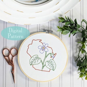Può includere: Arte di ricamo a cerchio con un fiore di viola viola con foglie verdi e un contorno marrone dello stato del Wisconsin. Il cerchio è su una superficie bianca con un paio di forbici e una pianta verde.