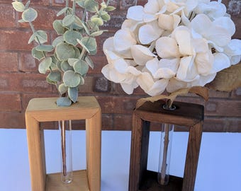 May include: Two rectangular wooden propagation stations with glass test tubes. One frame is light natural wood holding a green eucalyptus stem, and the other is dark stained wood holding a large, white, multi-petaled hydrangea flower head.