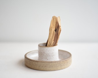 May include: A white ceramic incense holder with a speckled glaze. The holder has a small, round dish and a taller, cylindrical centre piece. A brown stick of incense is placed in the centre piece.