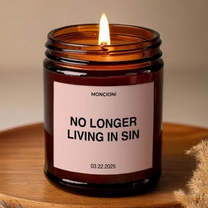 May include: A brown glass candle with a pink label that reads "NO LONGER LIVING IN SIN" and "MONCIONI" with the date "03.22.2025". The candle is lit and the flame is visible.