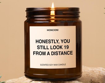 May include: A brown glass candle with a white label that reads "Honestly, you still look 19 from a distance." The candle is lit and sitting on a rock.