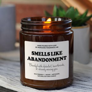 May include: A brown glass candle jar with a white label that reads "Smells Like Abandonment" and "Blended with disbelief, heartbreak, & already missing you." The label also includes the text "Hand Poured with Love by Moncioni Fragrance Lab" and "Eco Friendly | Vegan | Artisanal Natural Soy Wax Candle".