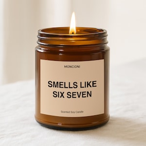 May include: A lit scented soy candle in a brown glass jar. The label reads "Smells Like Six Seven" with the brand name "Moncioni" above. The candle has a warm, inviting glow.