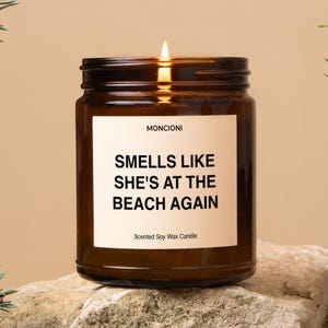 May include: A lit, brown glass jar candle with a white label. The label reads "Smells Like She's at the Beach Again" in black text. The candle is a scented soy wax candle.