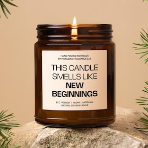 May include: A lit brown glass jar candle with a white label. The label reads "THIS CANDLE SMELLS LIKE NEW BEGINNINGS." The candle is on a rock surface. The candle is eco-friendly, vegan, artisanal, and made with natural soy wax.