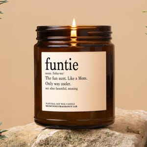 May include: A brown glass candle jar with a white label that reads "funtie" and a definition of the word. The candle is lit and the flame is visible. The candle is on a rock surface.