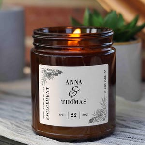 May include: A brown glass candle with a white label that reads "Congratulations on your ENGAGEMENT. ANNA & THOMAS. APRIL 22 2021." The label has a floral design.