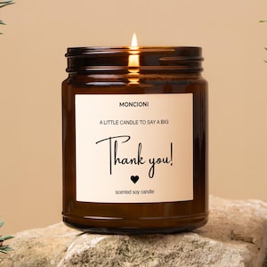 May include: A lit scented soy candle in a brown glass jar. The label reads "MONCIONI, A LITTLE CANDLE TO SAY A BIG Thank you!" with a heart symbol. The candle sits on a light-colored rock.