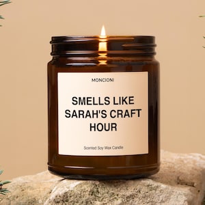 May include: A lit soy wax candle in a brown glass jar. The label reads "Smells Like Sarah's Craft Hour." The candle is on a stone surface. The candle is from the brand "Moncioni."