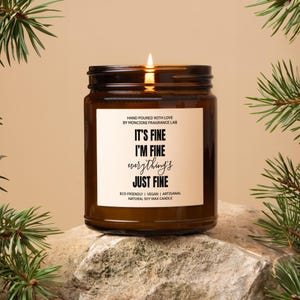 May include: A brown glass candle with a white label that reads "IT'S FINE I'M FINE everything's JUST FINE". The candle is hand-poured with love by Moncioni Fragrance Lab and is eco-friendly, vegan, and artisanal.