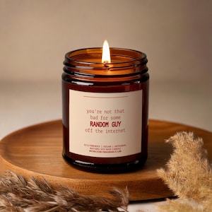 May include: A brown glass candle with a white label that reads "you're not that bad for some RANDOM GUY off the internet". The candle is lit and has a red and white wick. The label also includes the text "ECO FRIENDLY | VEGAN | ARTISANAL NATURAL SOY WAX CANDLE MONCIONI FRAGRANCE LAB".