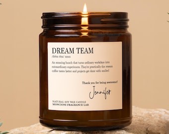 May include: A lit brown glass jar candle with the text "DREAM TEAM" on a white label. The label includes a definition and a thank you message. The candle is on a stone surface.